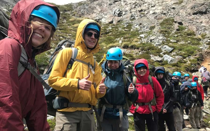 A group of people wearing safety gear and backpacks smile at the camera while hiking along a rocky trail.